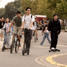 People on bikes, scooters, and skateboards navigate a busy traffic circle on the UC Davis campus with trees in the background.